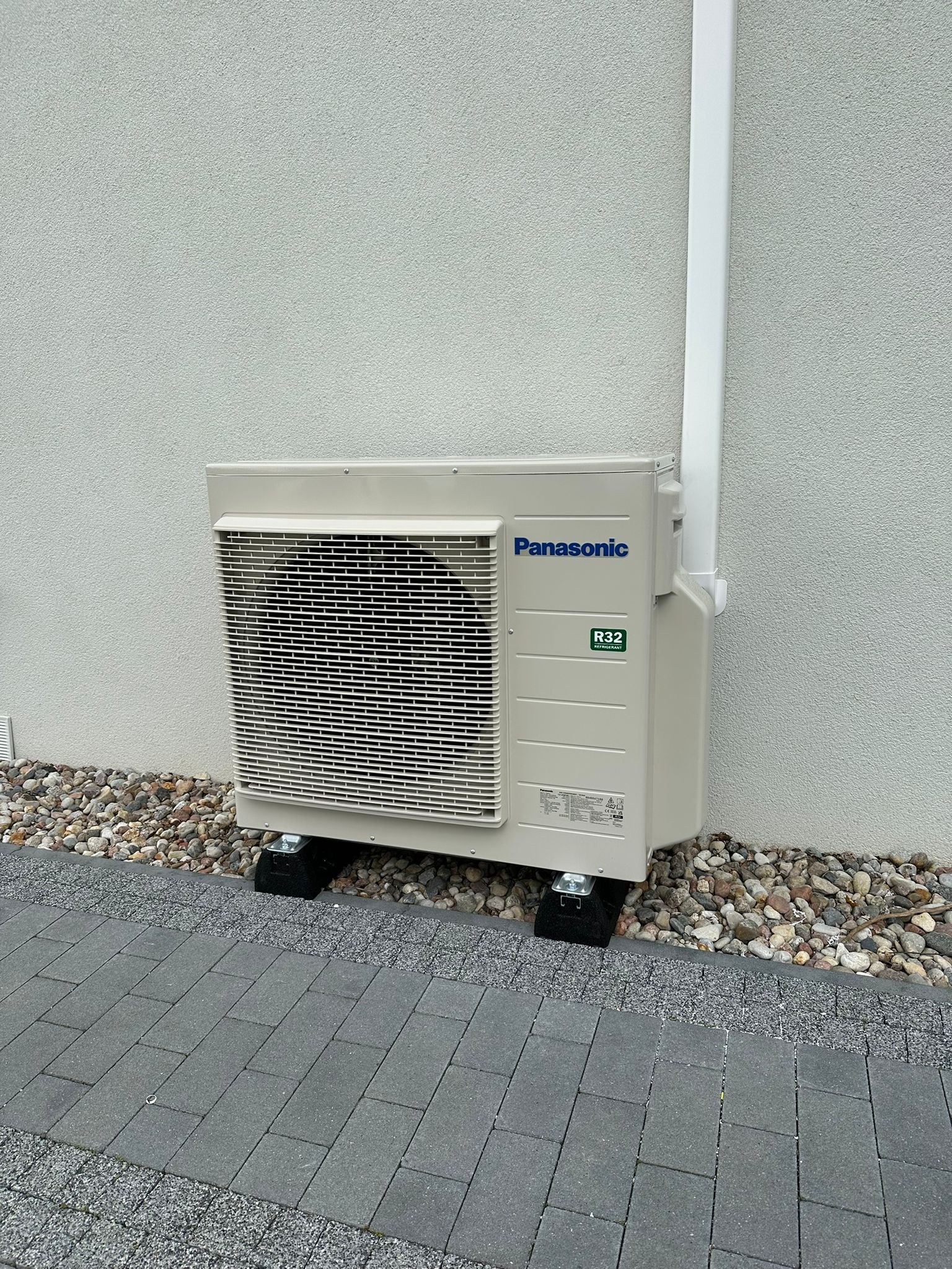Zamontowana jednostka zewnętrzna klimatyzacji Panasonic R32 na podkładkach, przy ścianie z kamieniami i rurą spustową. Widok z boku na kostce brukowej.