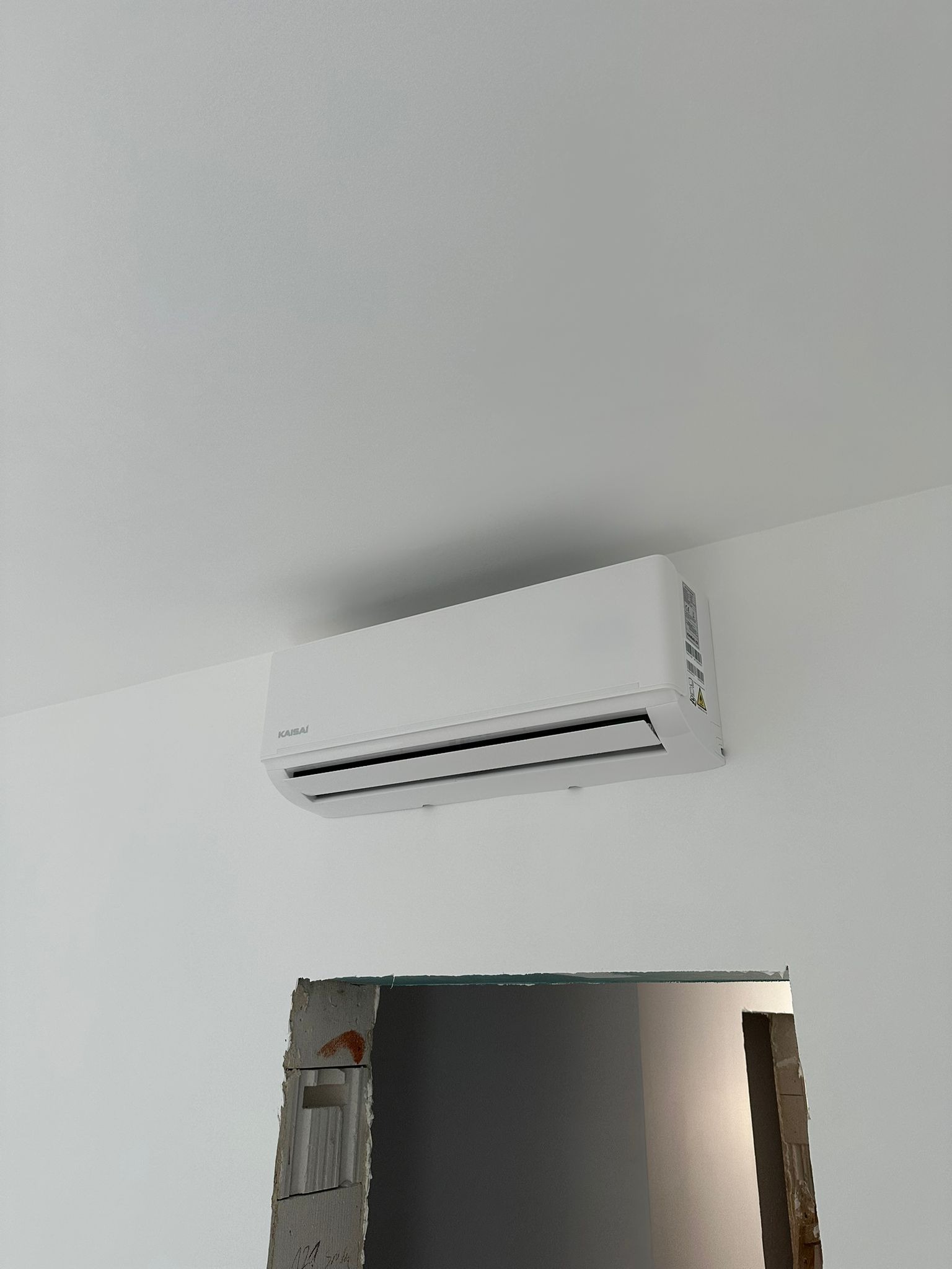 Biały klimatyzator Kaisai na białej ścianie, widoczny fragment otworu w ścianie, sufit w tle. Minimalistyczny design, oświetlenie naturalne.