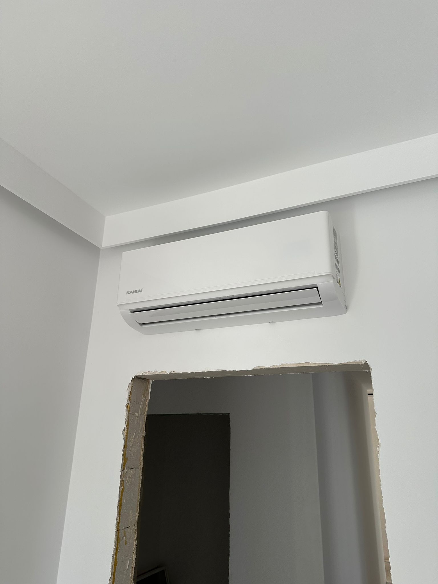 Biała klimatyzacja Kaisai w nowym, pomalowanym wnętrzu z widocznym otworem w ścianie. Minimalistyczny design urządzenia w jasnym pomieszczeniu.