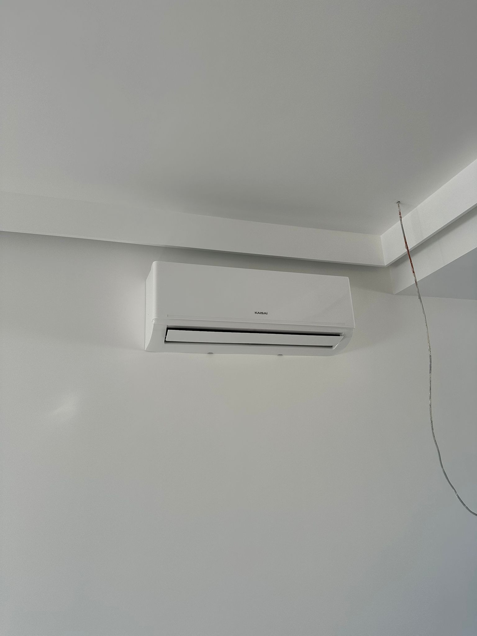 Biała klimatyzacja Kaisa montowana na ścianie w jasnym pomieszczeniu. Widoczne wystające kable elektryczne, sugerujące świeży montaż.