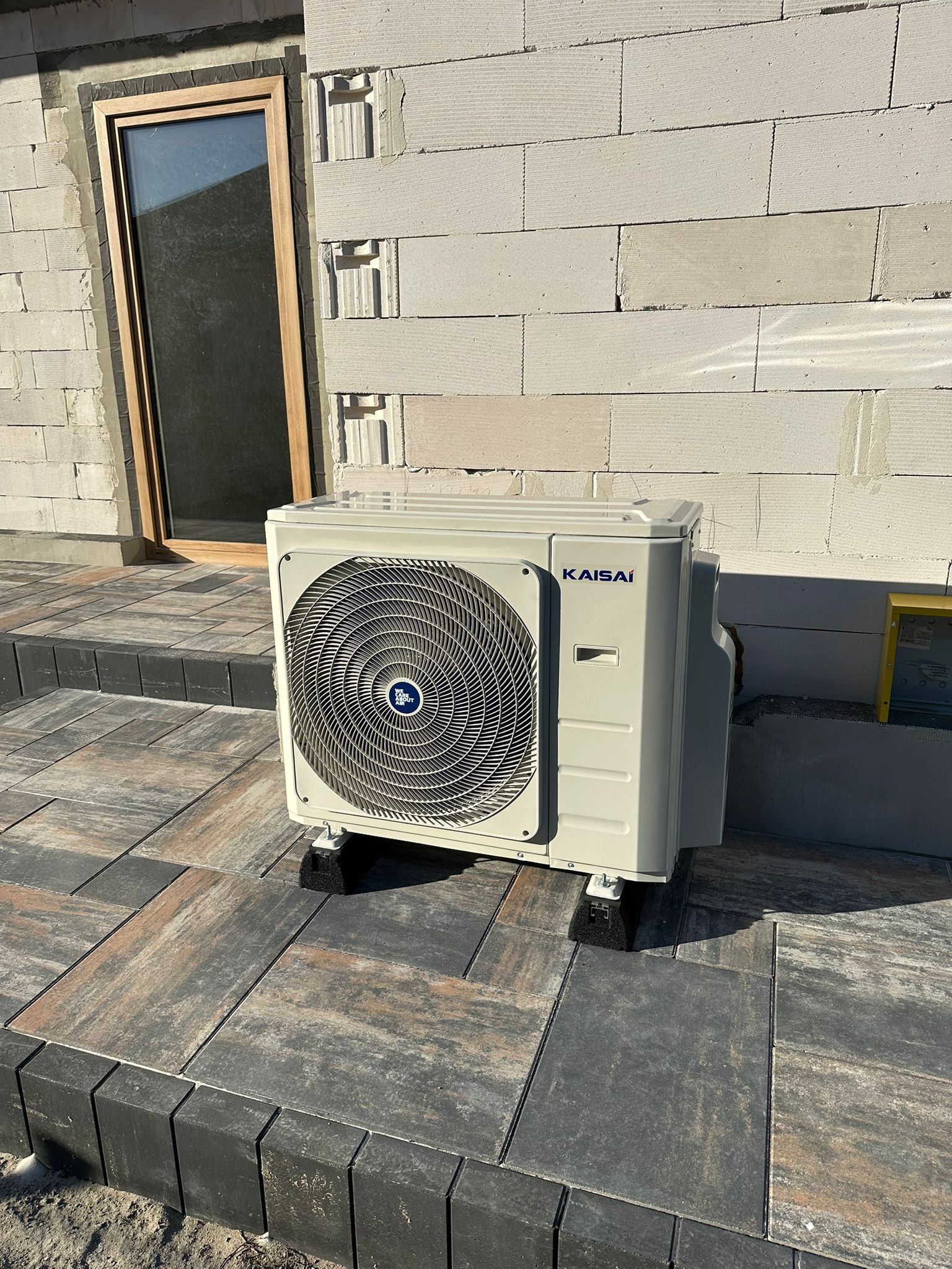 Zewnętrzna jednostka klimatyzacji Kaisai zamontowana na kostce brukowej przy ścianie budynku z betonu komórkowego. Widoczne okno z drewnianą ramą.