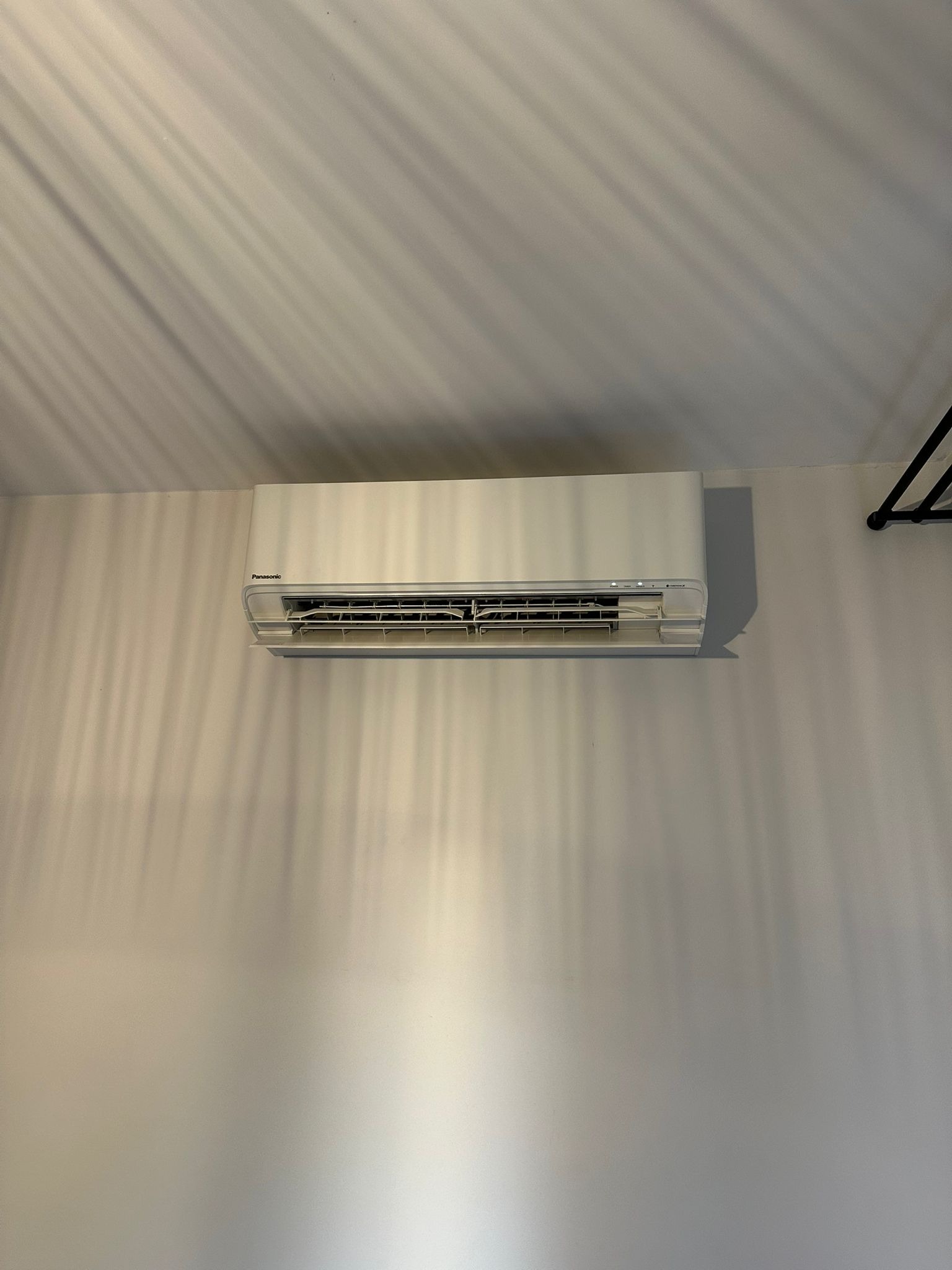 Biały klimatyzator Panasonic zamontowany na ścianie z efektem cienia, z widocznym fragmentem czarnego wieszaka w prawym górnym rogu.