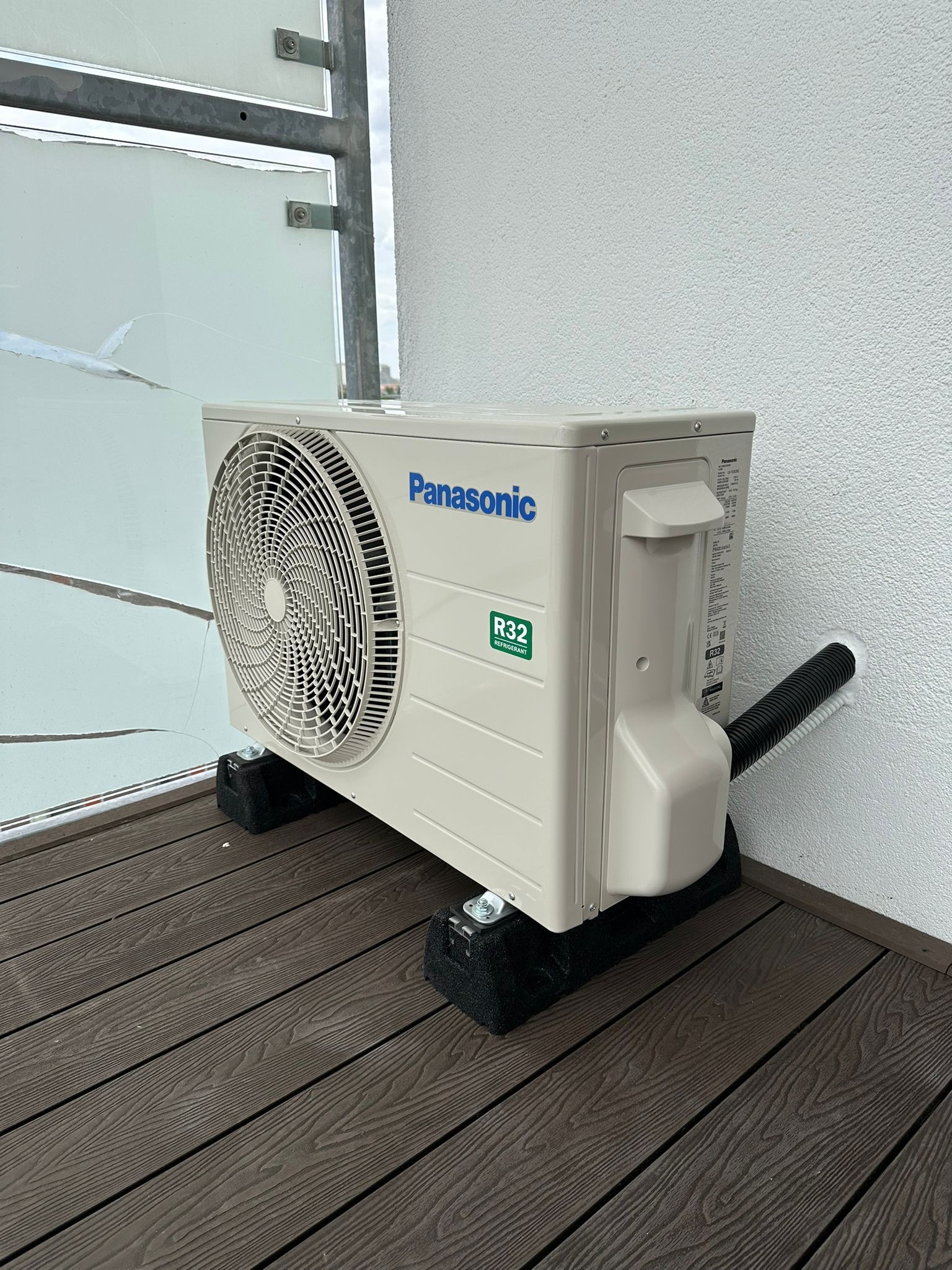 Biała jednostka zewnętrzna klimatyzacji Panasonic R32 na balkonie z drewnianą podłogą. Widoczne przewody i wsporniki. Ściana biała, tło stanowi szyba balkonu.