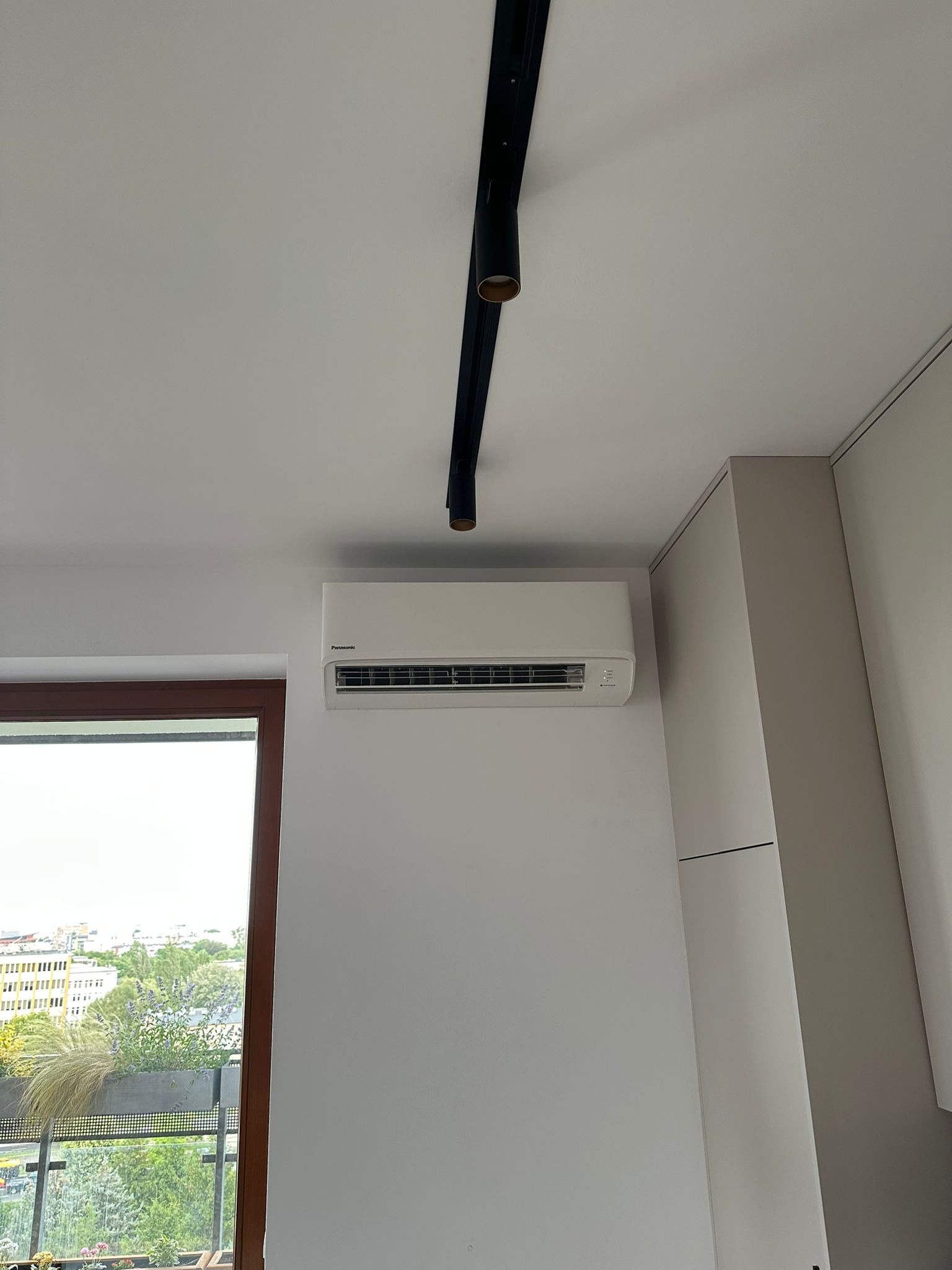 Biały klimatyzator Panasonic na ścianie w jasnym wnętrzu, z widokiem na miasto przez okno. Czarny reflektor sufitowy dodaje nowoczesnego charakteru.