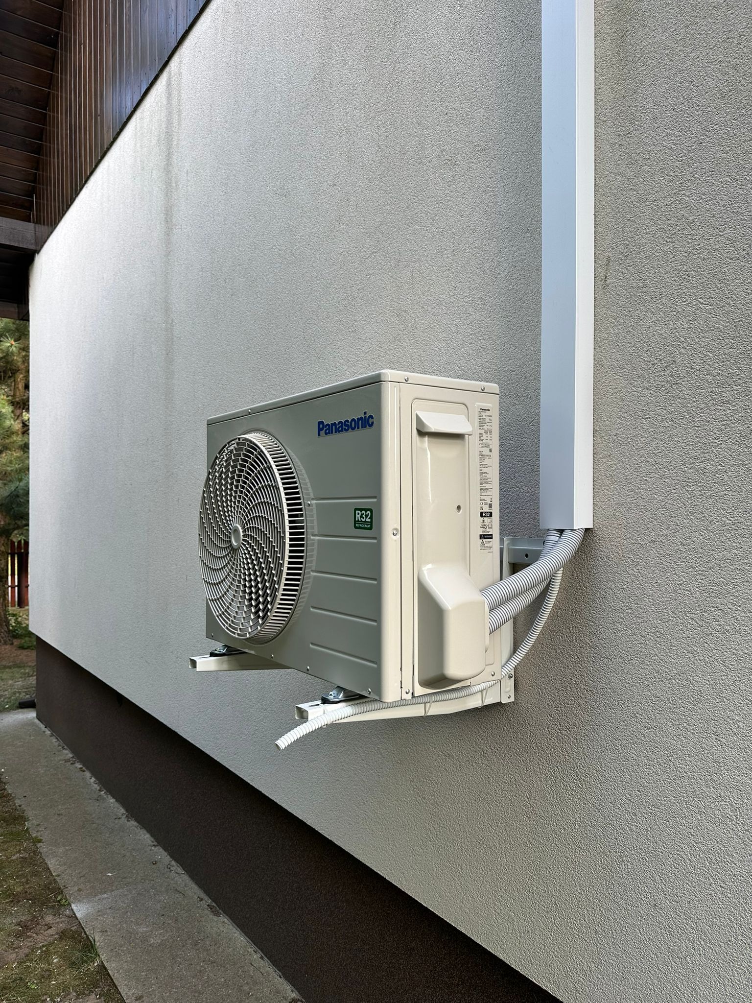 Zamontowana jednostka zewnętrzna klimatyzacji Panasonic R32 na elewacji budynku z widocznymi przewodami i osłoną. Szara ściana z fakturą tynku.