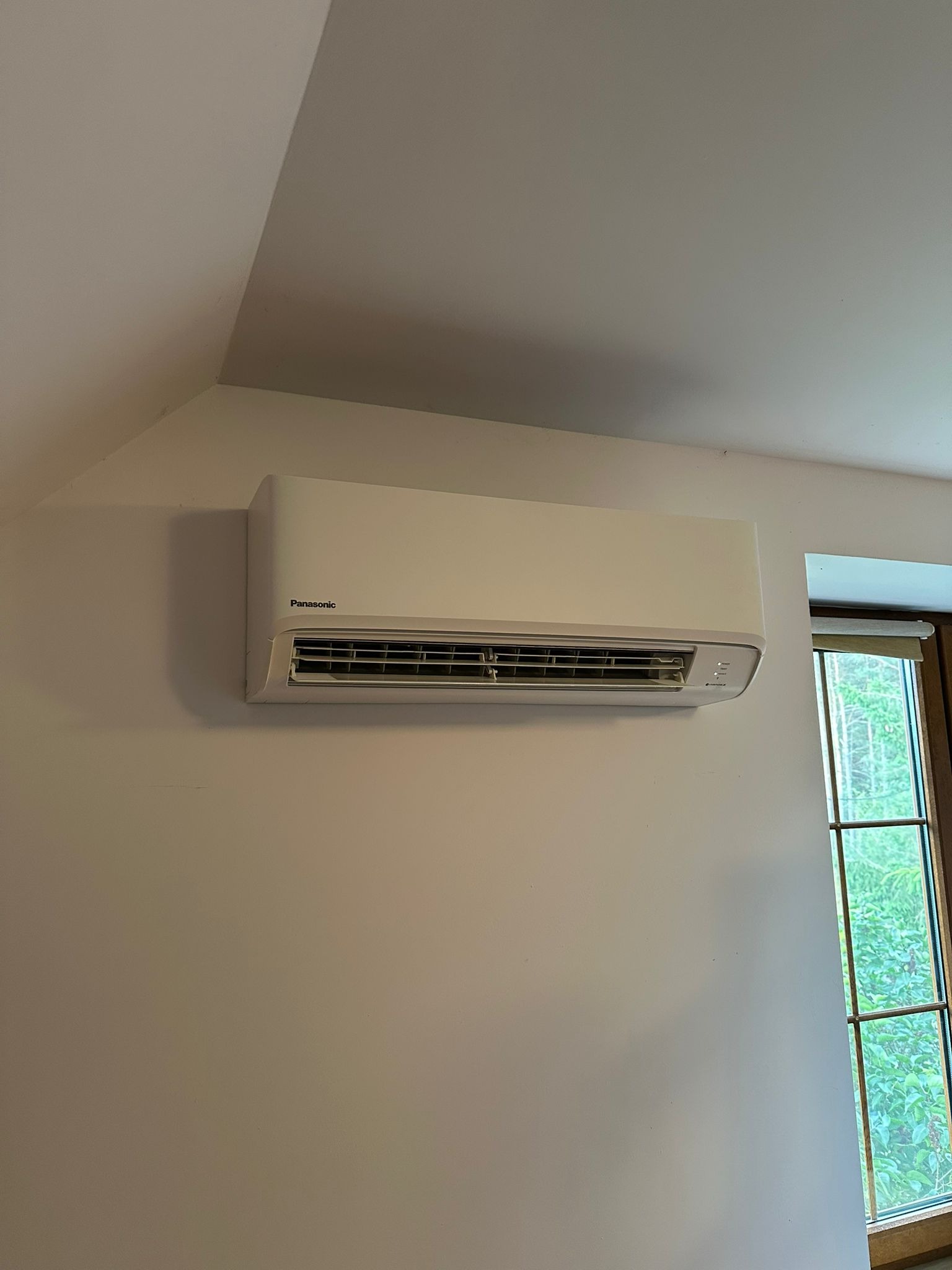 Biała, naścienna klimatyzacja Panasonic zamontowana w pokoju z oknem. Widoczny fragment skośnego sufitu.