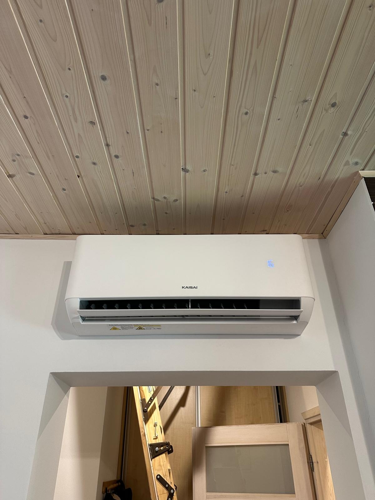 Biała klimatyzacja Kaisai zamontowana na ścianie w pomieszczeniu z drewnianym sufitem, widoczna z dołu pod lekkim kątem. W tle drzwi i drabina.