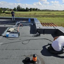 Prace dekarskie na płaskim dachu: montaż paneli solarnych i uszczelnianie kominka wentylacyjnego. Widoczne narzędzia, butla gazowa i maszyna do uszczelniania dachu.