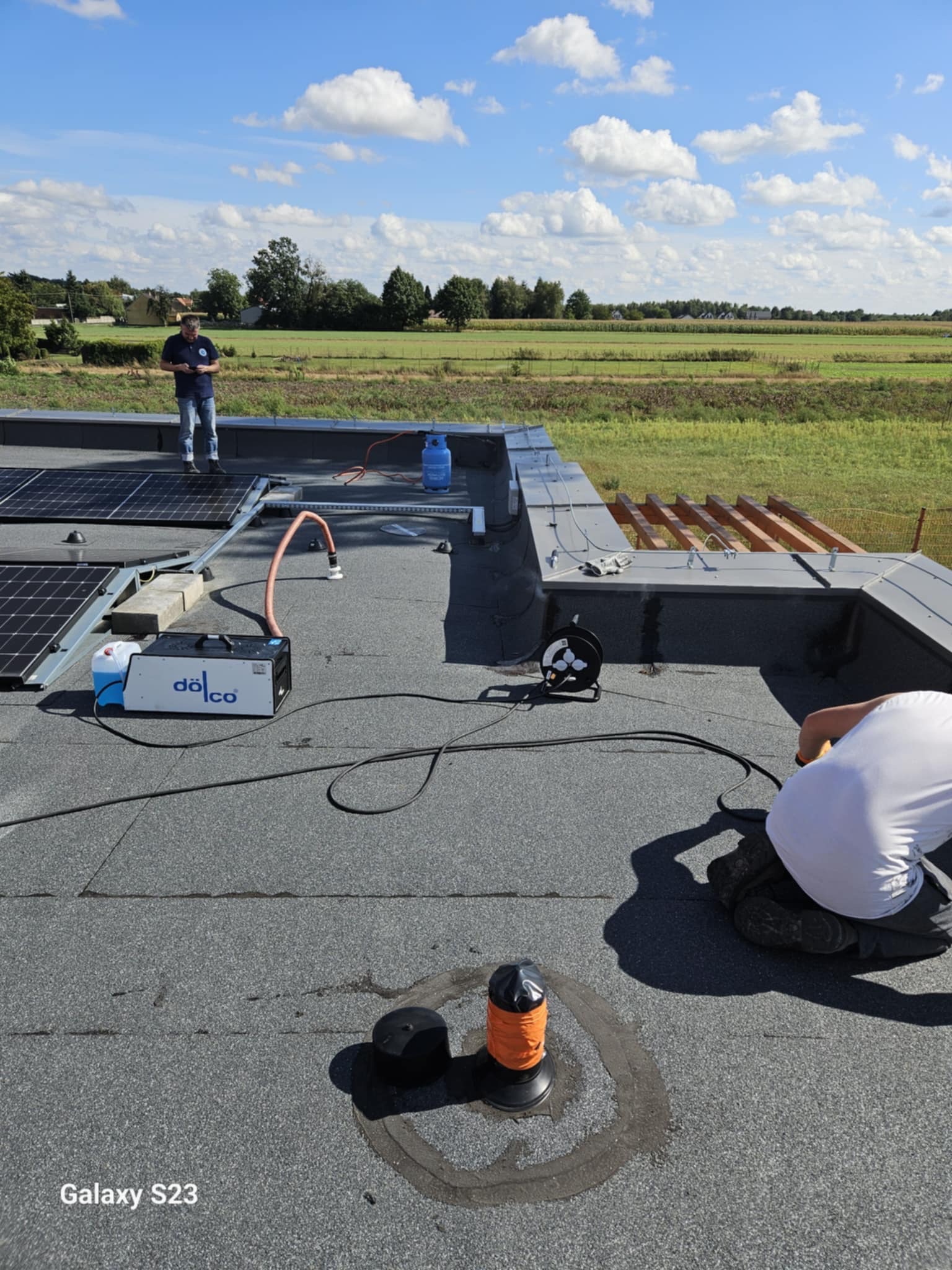 Prace dekarskie na płaskim dachu: montaż paneli solarnych i uszczelnianie kominka wentylacyjnego. Widoczne narzędzia, butla gazowa i maszyna do uszczelniania dachu.