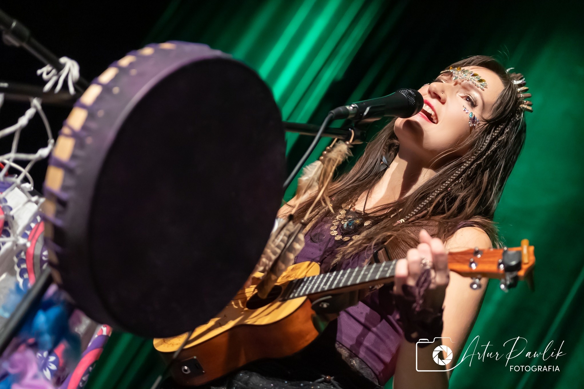 Artystka w fioletowej sukience śpiewa do mikrofonu, grając na ukulele. W tle zielona kurtyna i bęben szamański. Makijaż sceniczny i ozdoby na twarzy.