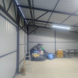 Michał Zbrzeźniak - Wnętrze hali z metalową konstrukcją i oświetleniem LED. Widoczne ściany z blachy trapezowej, drabina, zwinięty przedłużacz i sterta worków oraz kartonów.