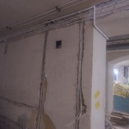 Michał Zbrzeźniak - Instalacja elektryczna w trakcie montażu: białe kable prowadzone po ścianie i suficie, widoczne puszki instalacyjne i fragmenty niezabezpieczonych ścian. Prace remontowe w toku.