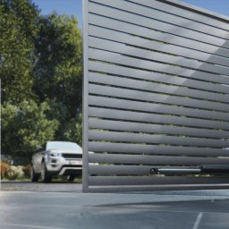 INTELIV MATEUSZ MUCHA - Nowoczesna, automatyczna brama przesuwna z szarych, poziomych paneli. Widoczny siłownik i częściowo otwarte skrzydło. W tle zaparkowany samochód i zieleń.