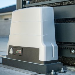 INTELIV MATEUSZ MUCHA - Szary napęd bramy przesuwnej Roger Brushless z listwą zębatą, zamontowany na kostce brukowej. Widoczny fragment domu w tle.