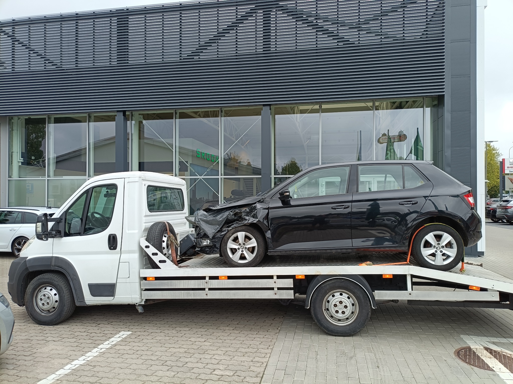 Biała laweta przewozi uszkodzony, czarny samochód marki Skoda na tle budynku salonu samochodowego Skoda z szarymi żaluzjami.