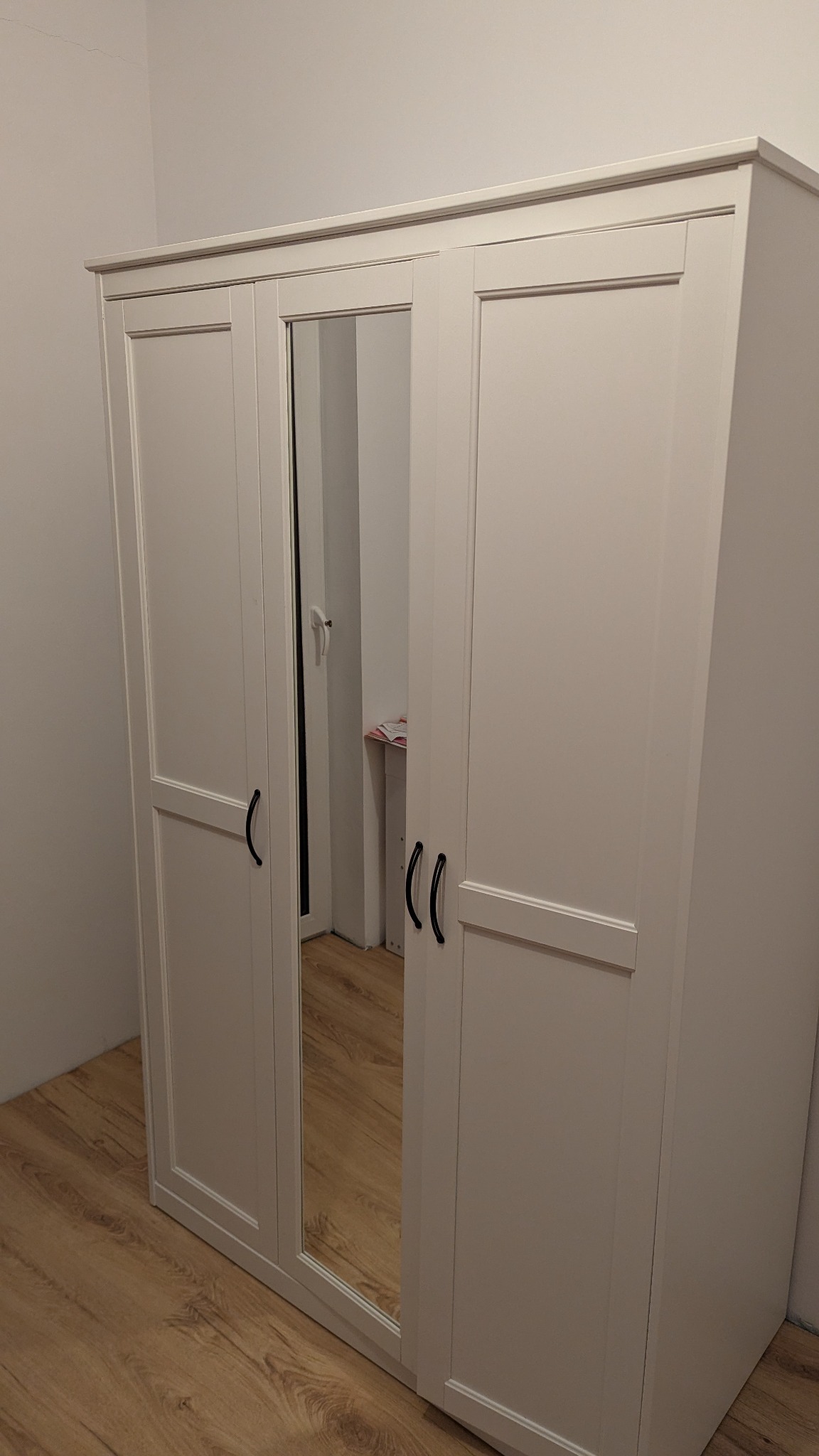 Trzydrzwiowa, biała szafa z lustrem pośrodku, z czarnymi uchwytami, ustawiona na drewnianej podłodze w rogu pokoju.