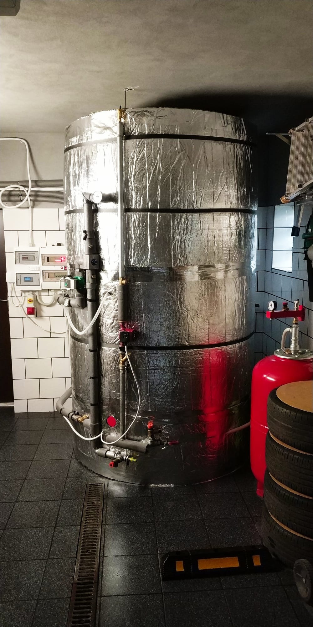 Duży, srebrny akumulator ciepła w kotłowni z białą glazurą i czarną podłogą. Widoczne rury, zawory i czerwony zbiornik wyrównawczy.