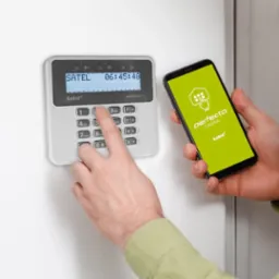 Panel sterowania alarmem Satel Perfecta z ekranem LCD, obsługiwany przez aplikację mobilną na smartfonie trzymanym w dłoni. Nowoczesny system alarmowy.
