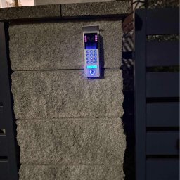MATEUSZ FORMELA ONECLICK - Nowoczesny wideodomofon z podświetlaną klawiaturą, zamontowany na kamiennym słupku ogrodzenia, widoczny fragment bramy. Oświetlenie nocne.