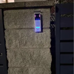 Nowoczesny wideodomofon z podświetlaną klawiaturą, zamontowany na kamiennym słupku ogrodzenia, widoczny fragment bramy. Oświetlenie nocne.