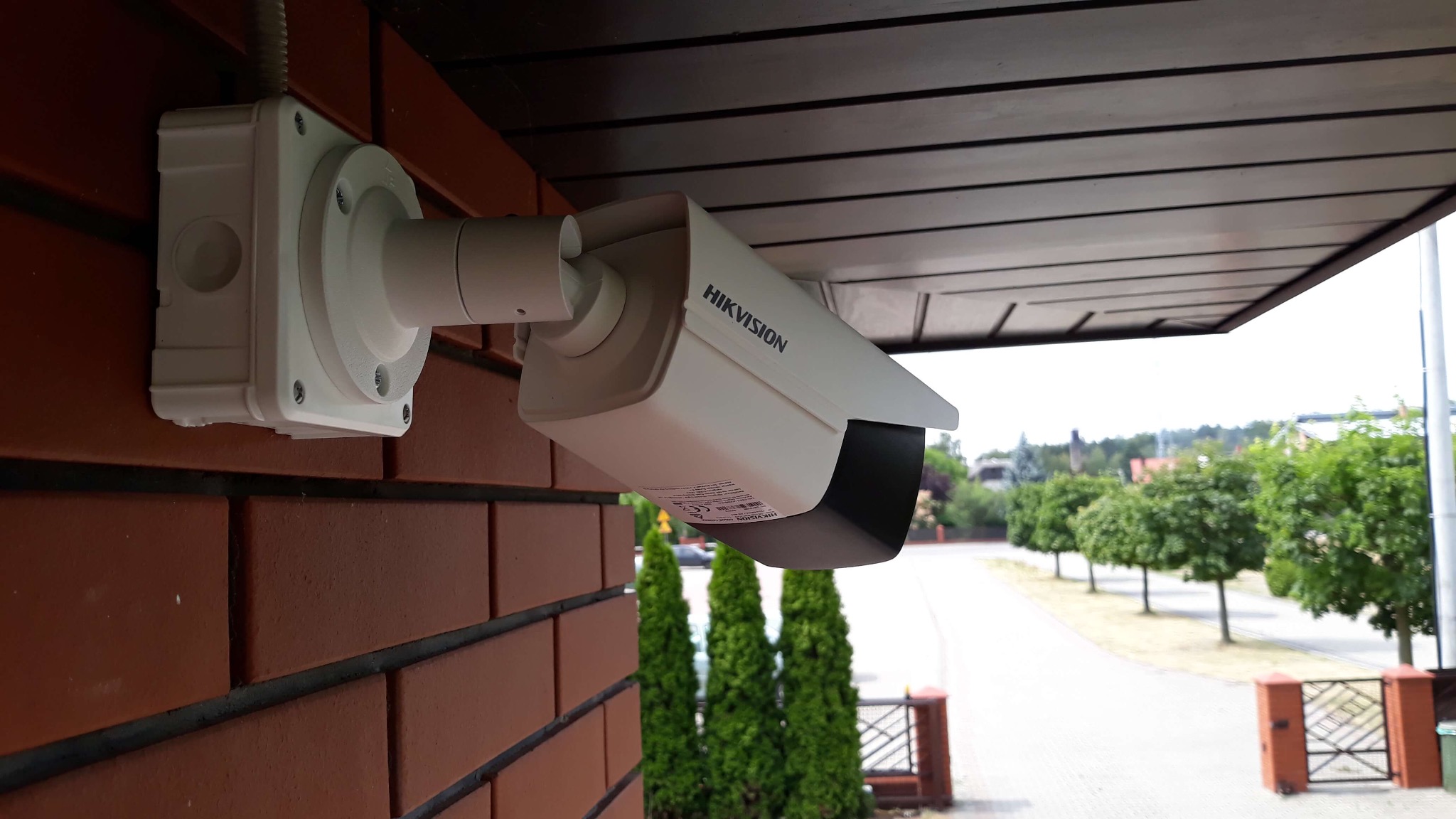Kamera monitoringu Hikvision zamontowana na ceglanej ścianie budynku, skierowana w stronę ulicy z zielenią. Widoczna skrzynka przyłączeniowa i fragment podsufitki.