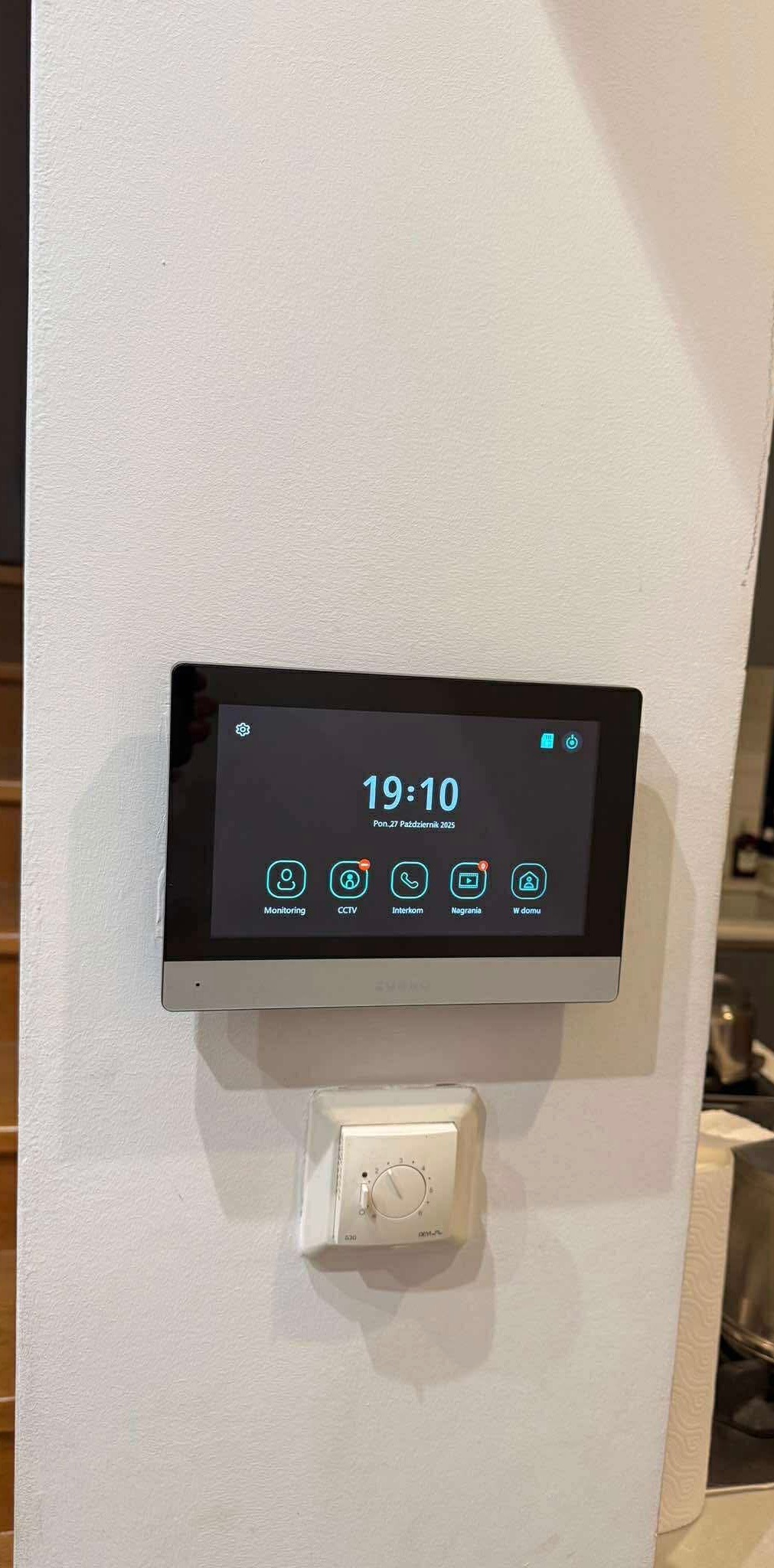 Nowoczesny panel dotykowy wideodomofonu z ikonami: monitoring, CCTV, interkom, nagrania, w domu. Zamontowany na ścianie, obok regulator temperatury.