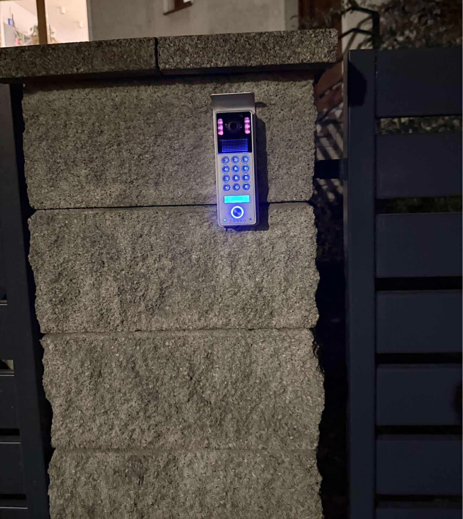 Nowoczesny wideodomofon z podświetlaną klawiaturą, zamontowany na kamiennym słupku ogrodzenia, widoczny fragment bramy. Oświetlenie nocne.