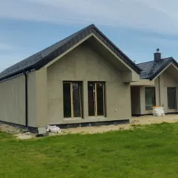 Nowoczesny dom jednorodzinny w stanie surowym zamkniętym z czarną dachówką i szarym tynkiem, widok z zewnątrz na tle błękitnego nieba i zielonego trawnika.