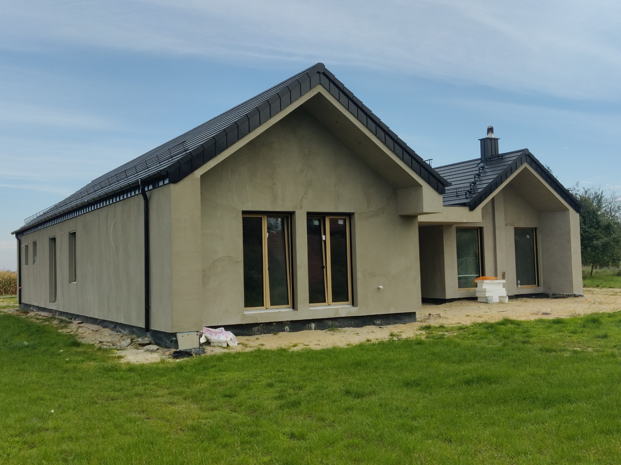 Nowoczesny dom jednorodzinny w stanie surowym zamkniętym z czarną dachówką i szarym tynkiem, widok z zewnątrz na tle błękitnego nieba i zielonego trawnika.