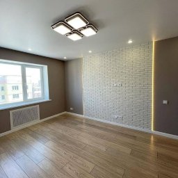 Prof&work Volodymyr Pasichnyk - Jasne wnętrze z drewnianą podłogą i ścianą z białej cegły, oświetloną listwą LED. Nowoczesna lampa sufitowa. Okno z widokiem na budynek mieszkalny.