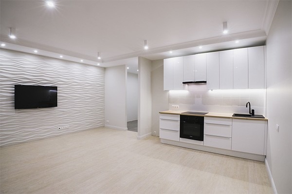 Nowoczesna, jasna kuchnia z białymi błyszczącymi szafkami i falistą ścianą z telewizorem. Minimalistyczny design, oświetlenie LED pod szafkami i punktowe na suficie.