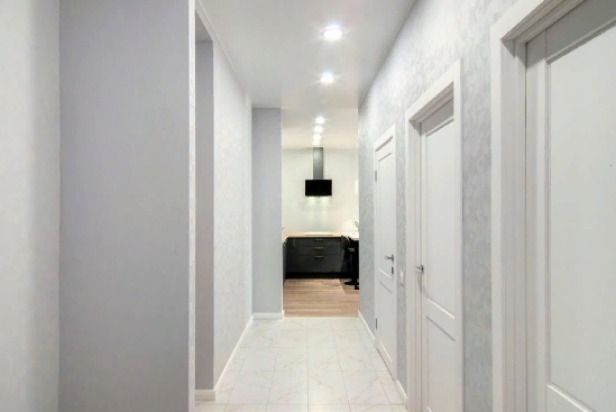 Wyremontowany korytarz z trzema białymi drzwiami i widokiem na kuchnię w tle, Warszawa. Ściany w odcieniach szarości z delikatnym wzorem, jasne płytki na podłodze.