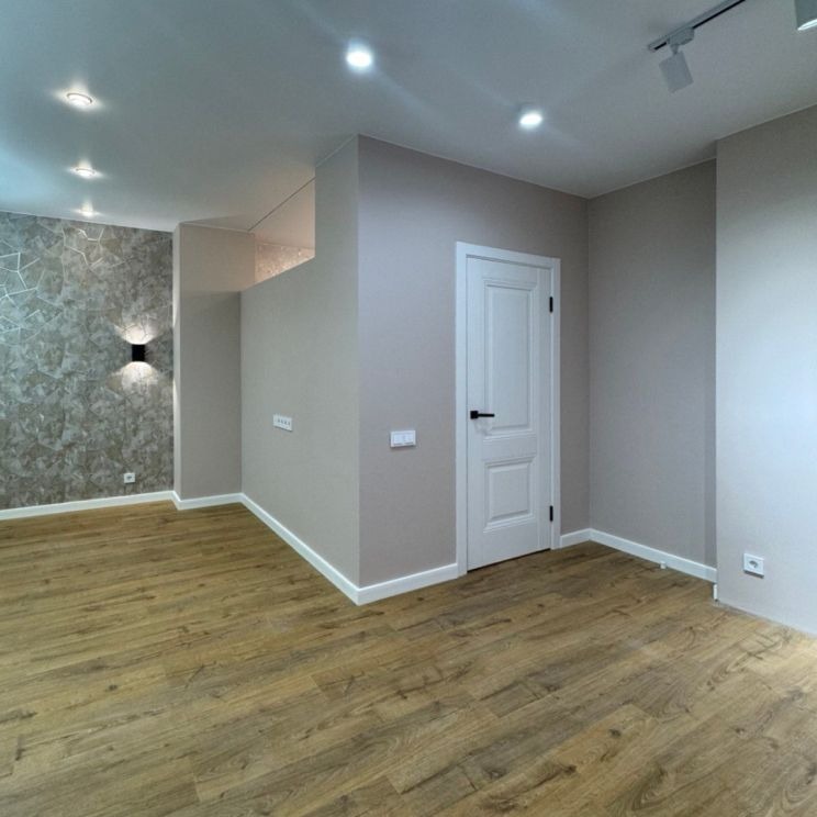 Jasne wnętrze po remoncie: drewnopodobna podłoga, beżowe ściany z dekoracyjną tapetą i białe drzwi z czarną klamką. Oświetlenie punktowe.