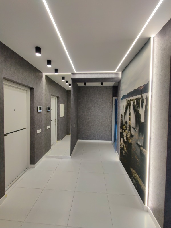 Nowoczesny korytarz z szarą tapetą, białymi drzwiami i podłogą. Oświetlenie LED w suficie i pionowe przy ścianie z fototapetą z motywem jeziora.