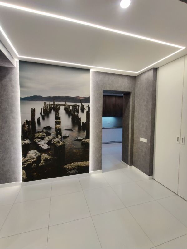 Nowoczesne wnętrze z fototapetą przedstawiającą krajobraz morski, oświetleniem LED w suficie i białymi płytkami podłogowymi. Widoczne drzwi do innego pomieszczenia.