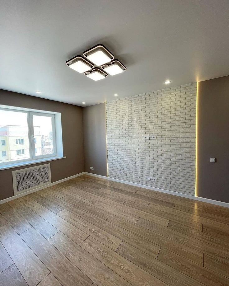 Jasne wnętrze z drewnianą podłogą i ścianą z białej cegły, oświetloną listwą LED. Nowoczesna lampa sufitowa. Okno z widokiem na budynek mieszkalny.
