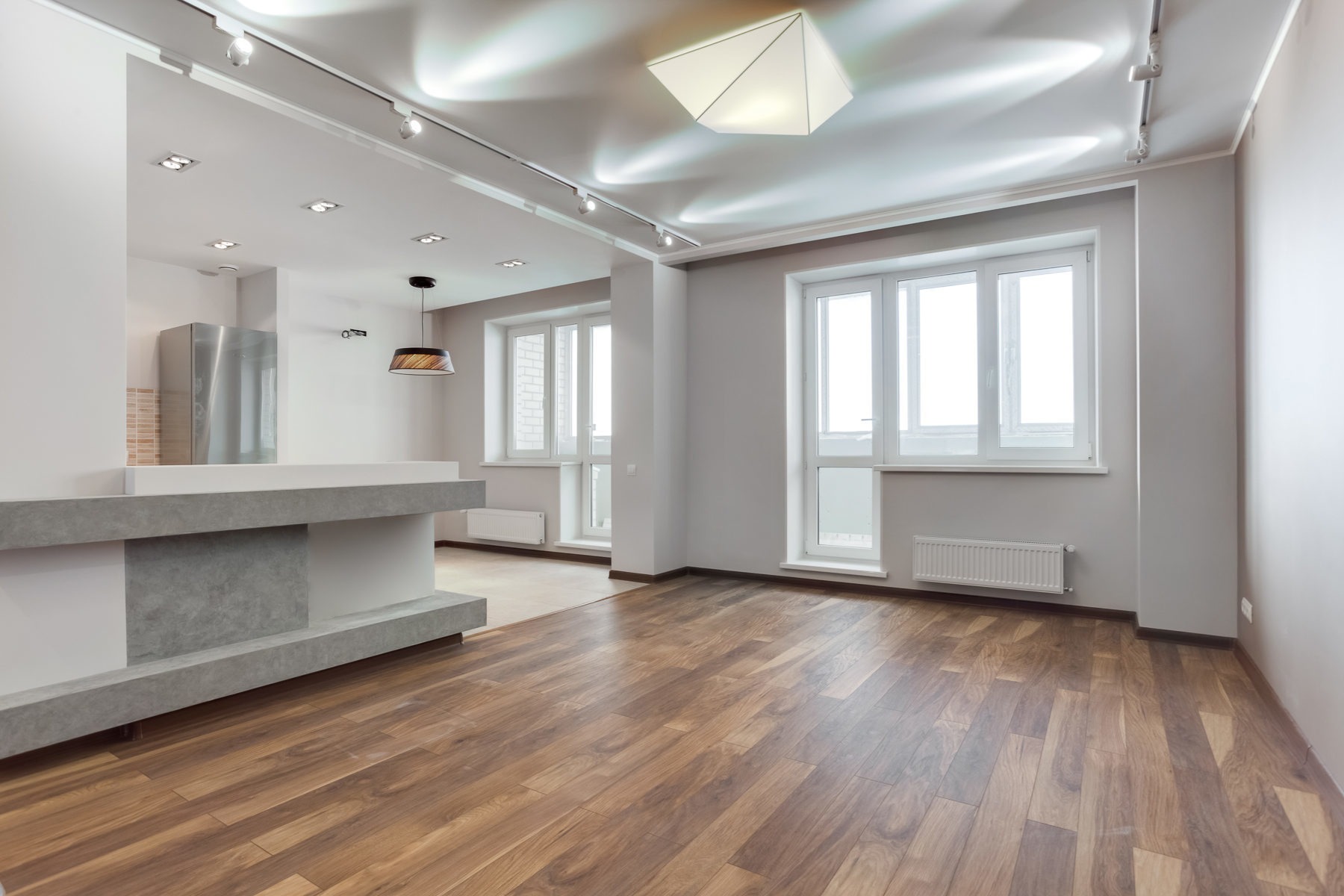 Jasne, przestronne wnętrze po remoncie: drewniana podłoga, szare ściany, białe okna i grzejniki. Widoczna część kuchni z blatem i nowoczesnym oświetleniem.