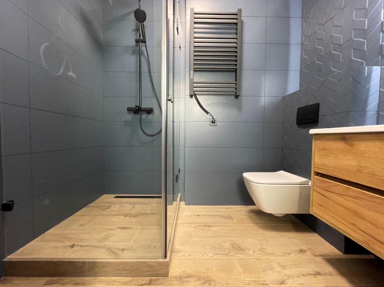 Nowoczesna łazienka w odcieniach szarości z drewnianymi akcentami. Kabina prysznicowa typu walk-in, podwieszana toaleta i grzejnik drabinkowy.