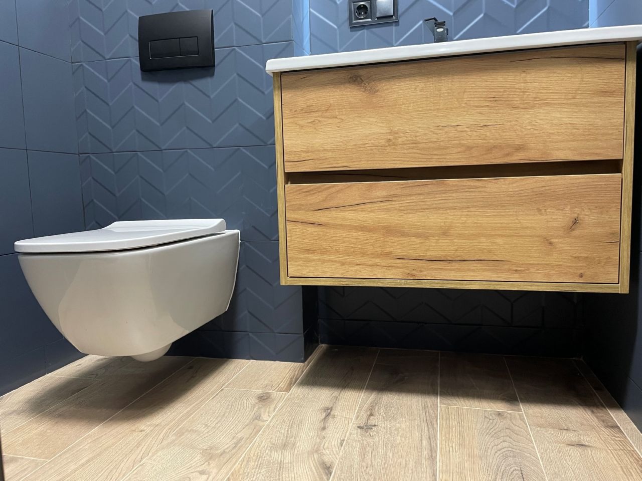 Nowoczesna łazienka: podwieszana toaleta, szafka z umywalką o drewnianym wykończeniu i granatowe płytki w jodełkę. Jasna drewniana podłoga dodaje ciepła.