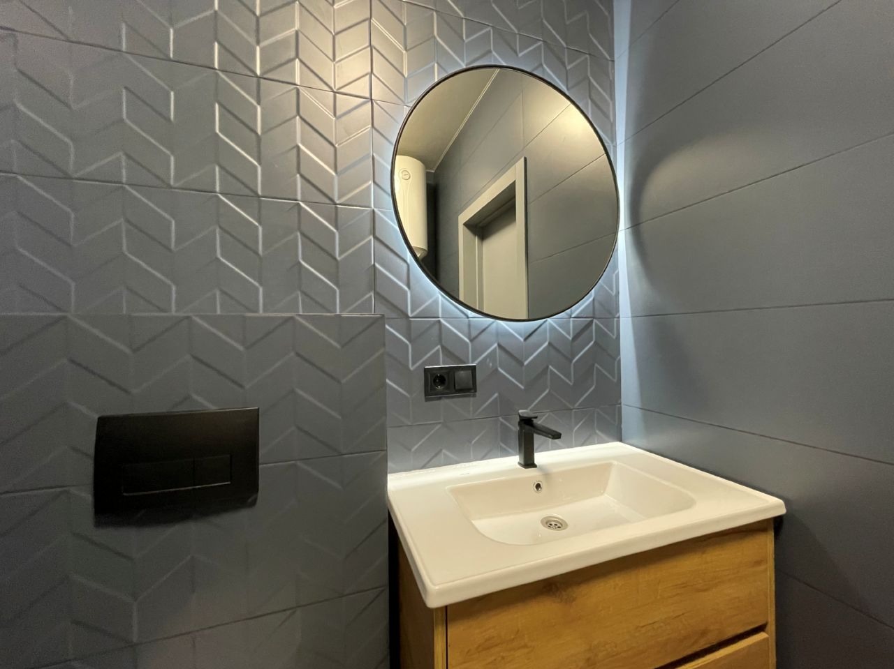Nowoczesna łazienka: szare płytki z wzorem jodełki, okrągłe lustro z podświetleniem LED, drewniana szafka pod umywalką i czarna armatura.