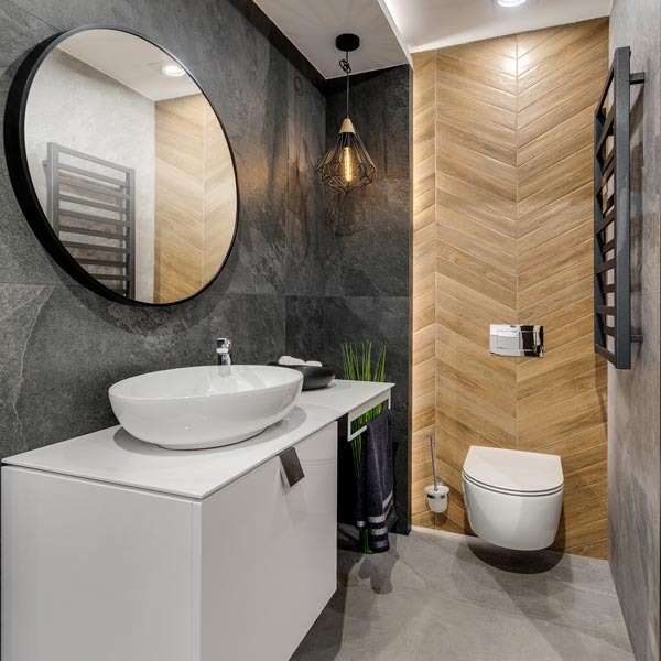 Nowoczesna łazienka z owalnym lustrem, białym blatem umywalki i drewnopodobnymi płytkami ułożonymi w jodełkę na ścianie. Czarny grzejnik drabinkowy.