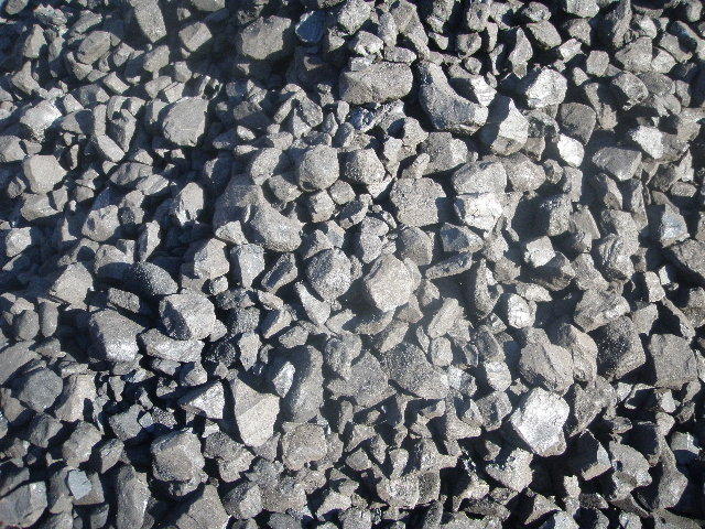 Kupa czarnych bryłek węgla brunatnego o nieregularnych kształtach, oświetlona jasnym światłem słonecznym, ukazująca strukturę materiału.