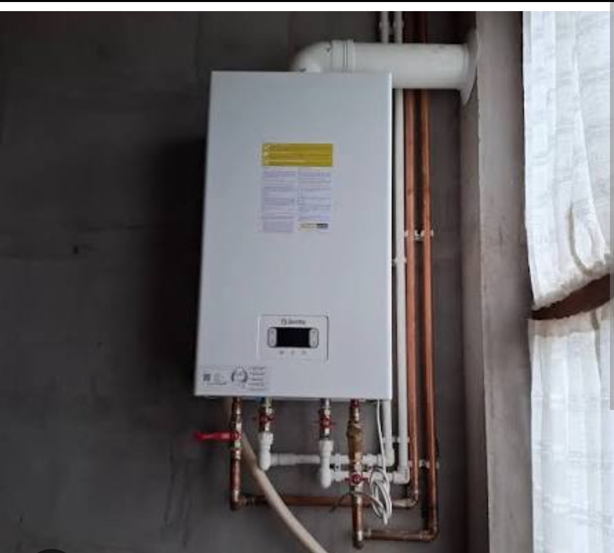 Biały piec gazowy marki Beretta, zamontowany na szarej ścianie, z widocznymi miedzianymi rurami instalacji. Obok okno z białą firanką. Ujęcie z dołu.