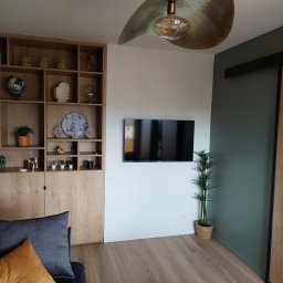 Artur Żurek - Nowoczesny salon z drewnianą zabudową, telewizorem na ścianie i designerską lampą. Parkiet w odcieniach brązu, zielona ściana i dekoracje dodają wnętrzu charakteru.