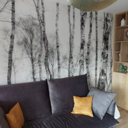 Artur Żurek - Stylowy salon z szarą sofą, złotymi poduszkami i fototapetą z motywem brzozowego lasu. Złota lampa sufitowa i drewniany regał dopełniają wystrój.