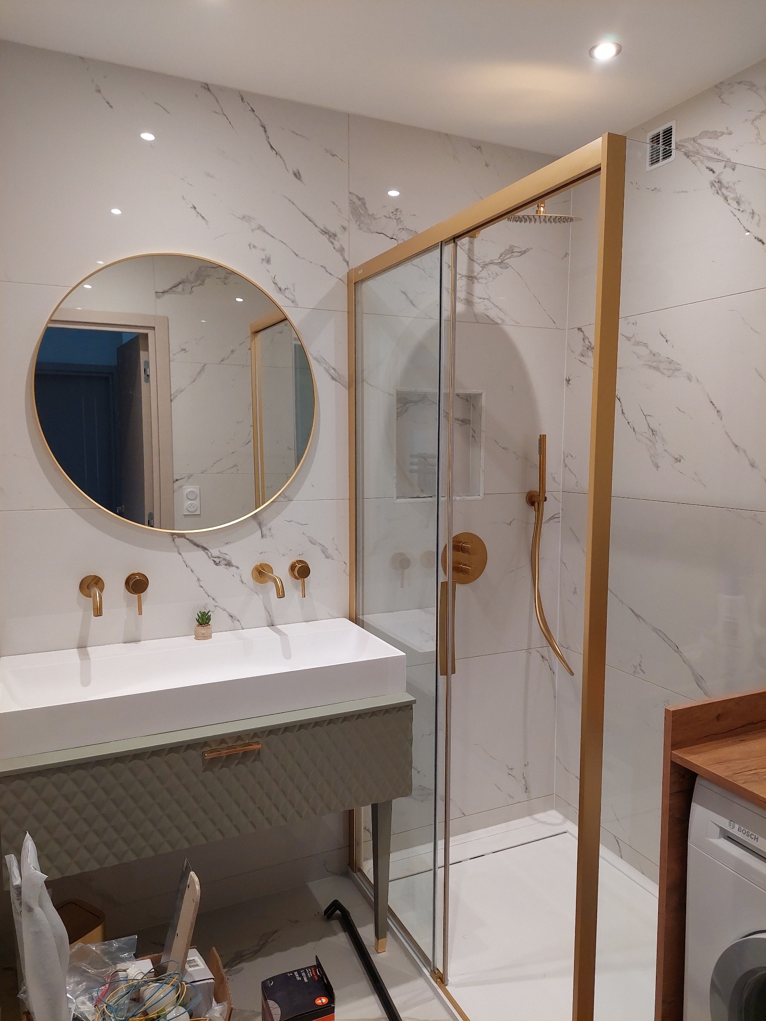 Nowoczesna łazienka z kabiną prysznicową w złotej ramie, umywalką z szafką w kolorze oliwkowym i okrągłym lustrem na tle białych płytek imitujących marmur.