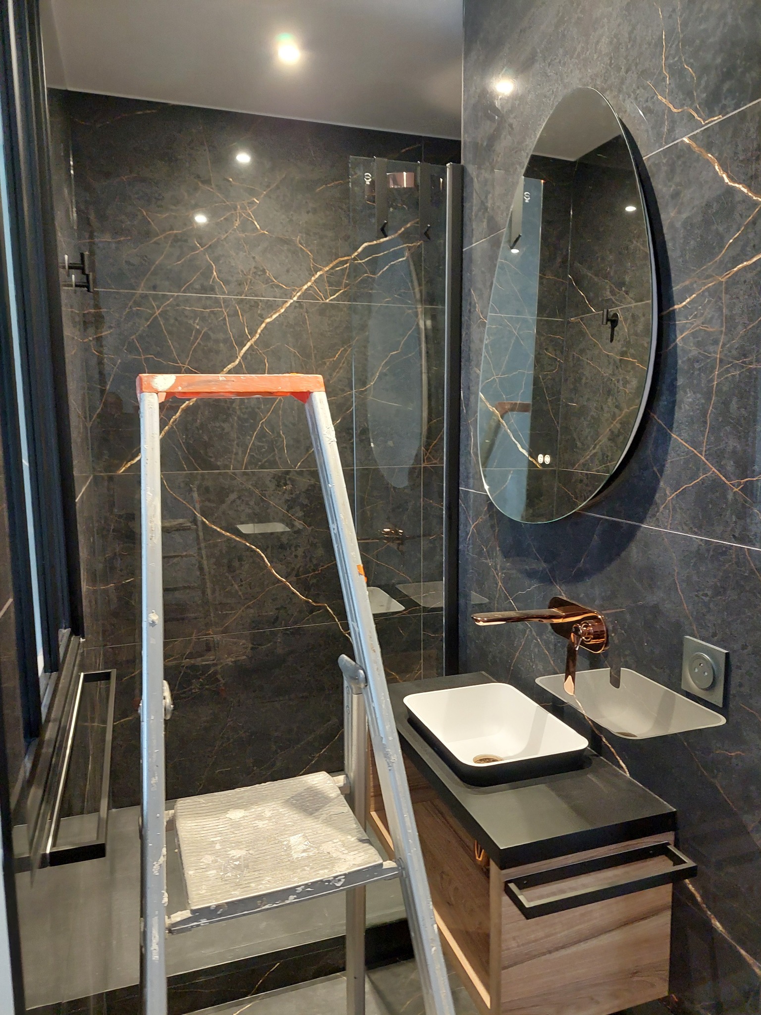 Nowoczesna łazienka z czarnymi płytkami imitującymi marmur, owalnym lustrem z podświetleniem i złotą armaturą. Widoczna drabina malarska sugeruje trwające prace wykończeniowe.