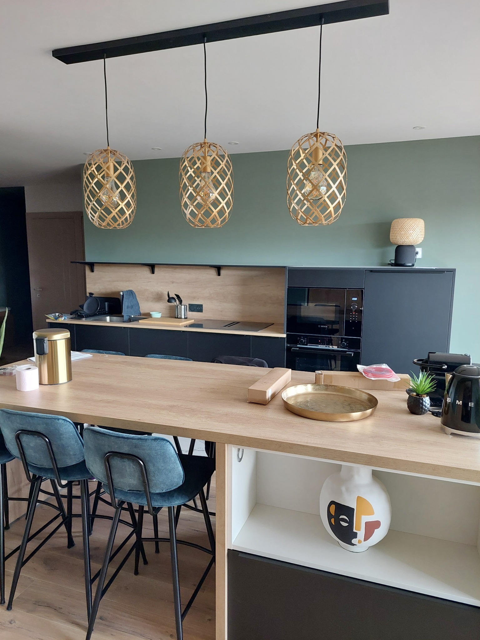 Nowoczesna kuchnia z wyspą i trzema ażurowymi lampami wiszącymi. Zielona ściana, czarne szafki i drewniany blat. Dekoracyjna ceramika i akcesoria na wyspie.