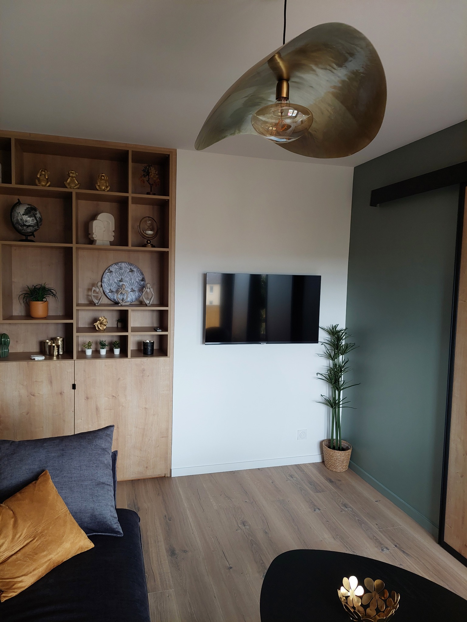 Nowoczesny salon z drewnianą zabudową, telewizorem na ścianie i designerską lampą. Parkiet w odcieniach brązu, zielona ściana i dekoracje dodają wnętrzu charakteru.