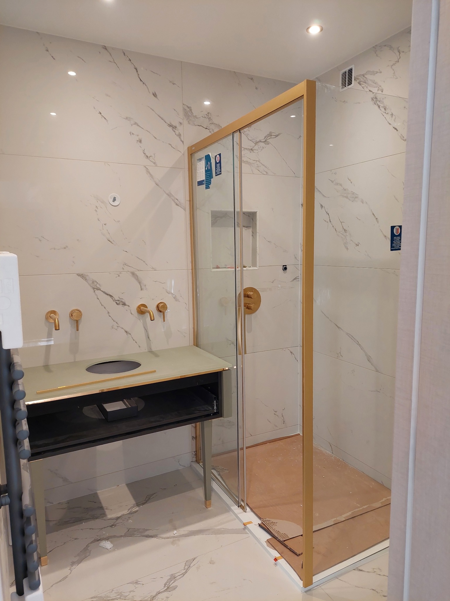 Nowoczesna łazienka w trakcie wykańczania: marmurowe płytki, złote akcenty armatury i kabiny prysznicowej, minimalistyczna umywalka. Widoczne zabezpieczenia podłogi w kabinie.